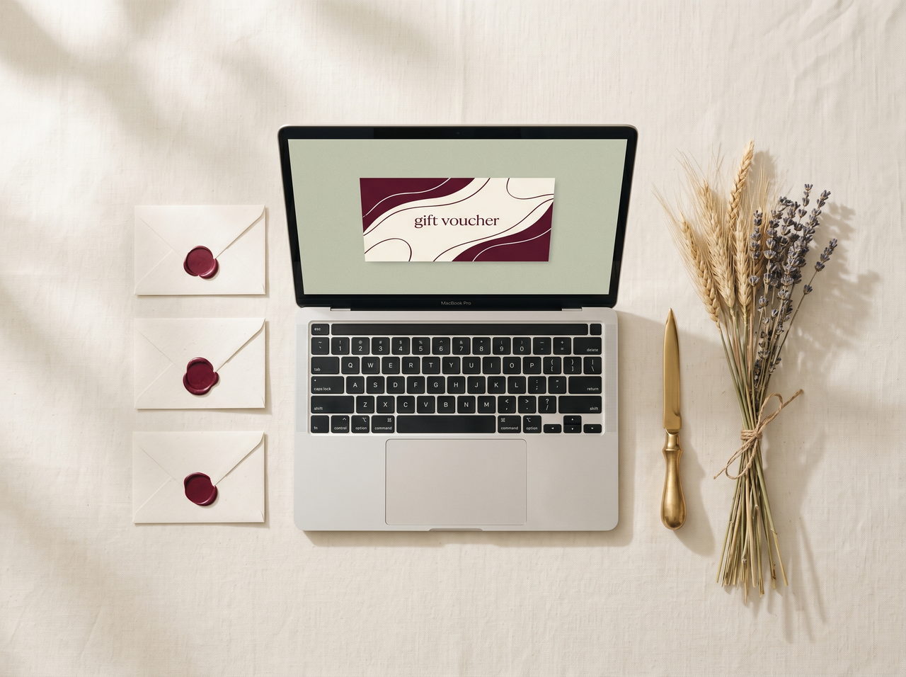 A laptop displaying a deep burgundy gift voucher beside three cream envelopes sealed with wax, dried wheat and lavender, and a brass letter opener on cream linen.